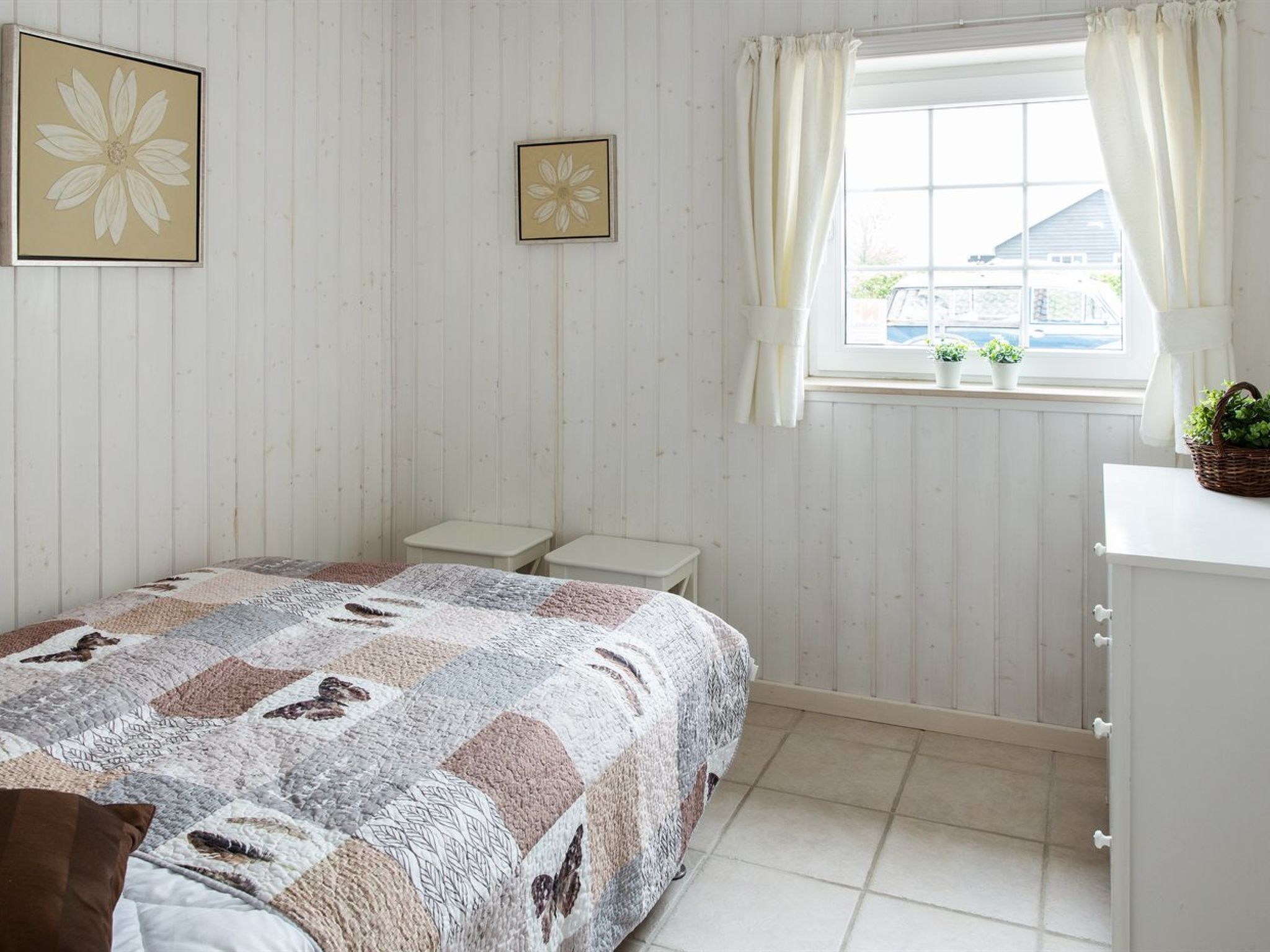 Ein Schlafzimmer mit einem Bett, Nachttischen, einem Fenster mit Vorhängen und zwei gerahmten Blumenbildern an der Wand.