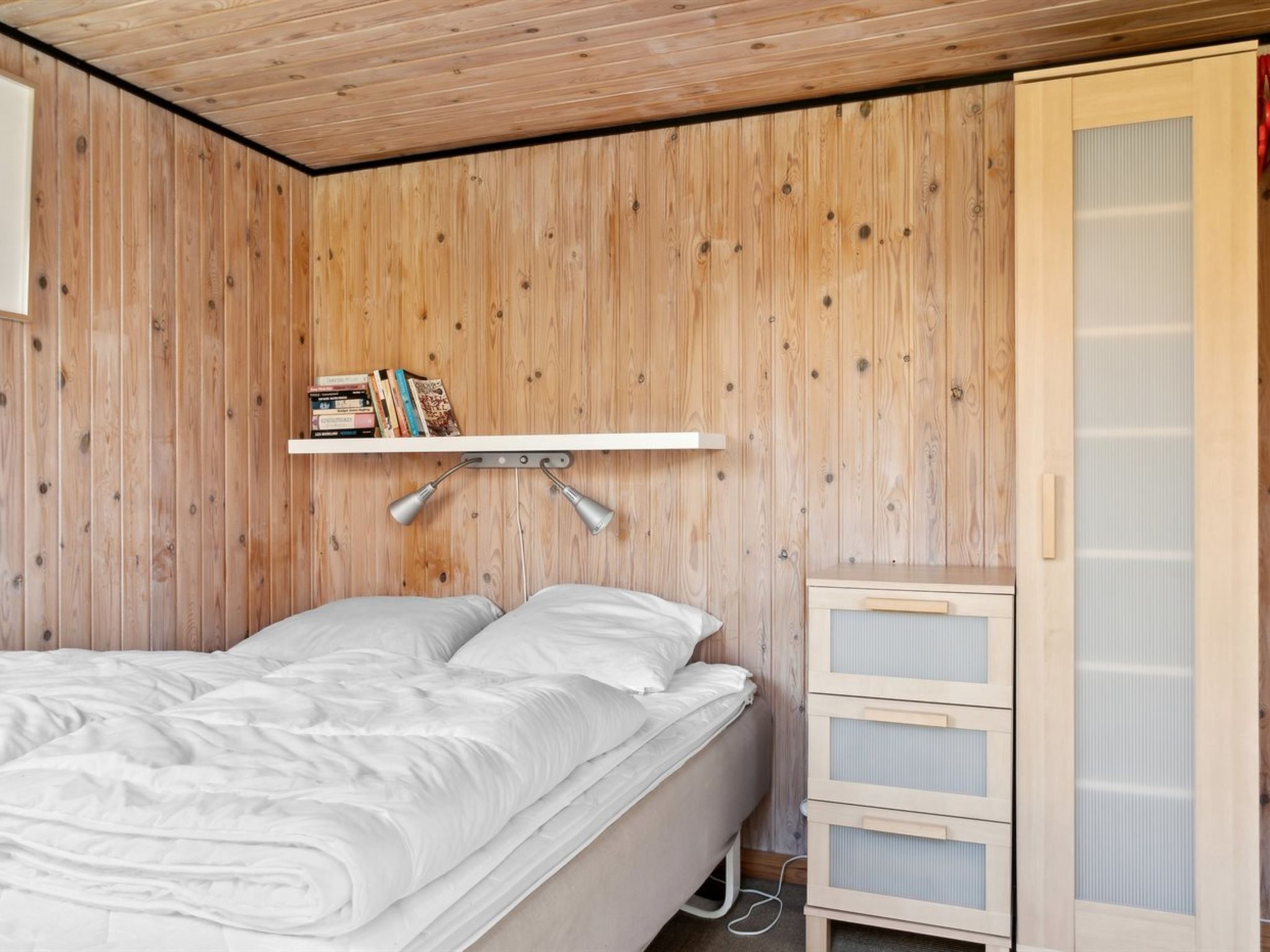 A bedroom with a wooden wall, a bed with white bedding, a shelf with books, and a wardrobe with a sliding door.
