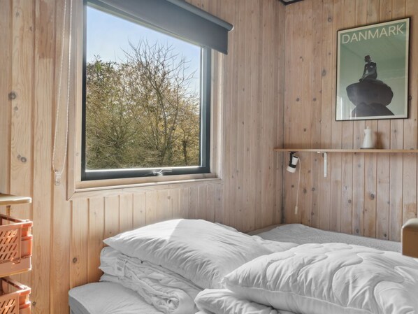 A bedroom with a large window, wooden walls, a bed with white bedding, a shelf with a Denmark poster, and a small table with a lamp.
