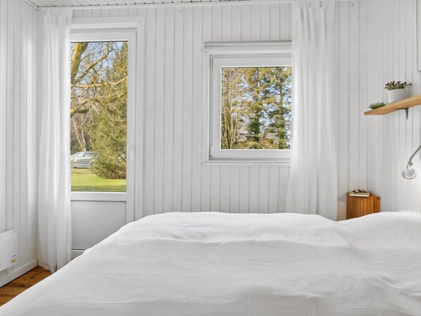 A bedroom with a large bed, two windows, a bedside table with a lamp, and a small shelf with a plant.