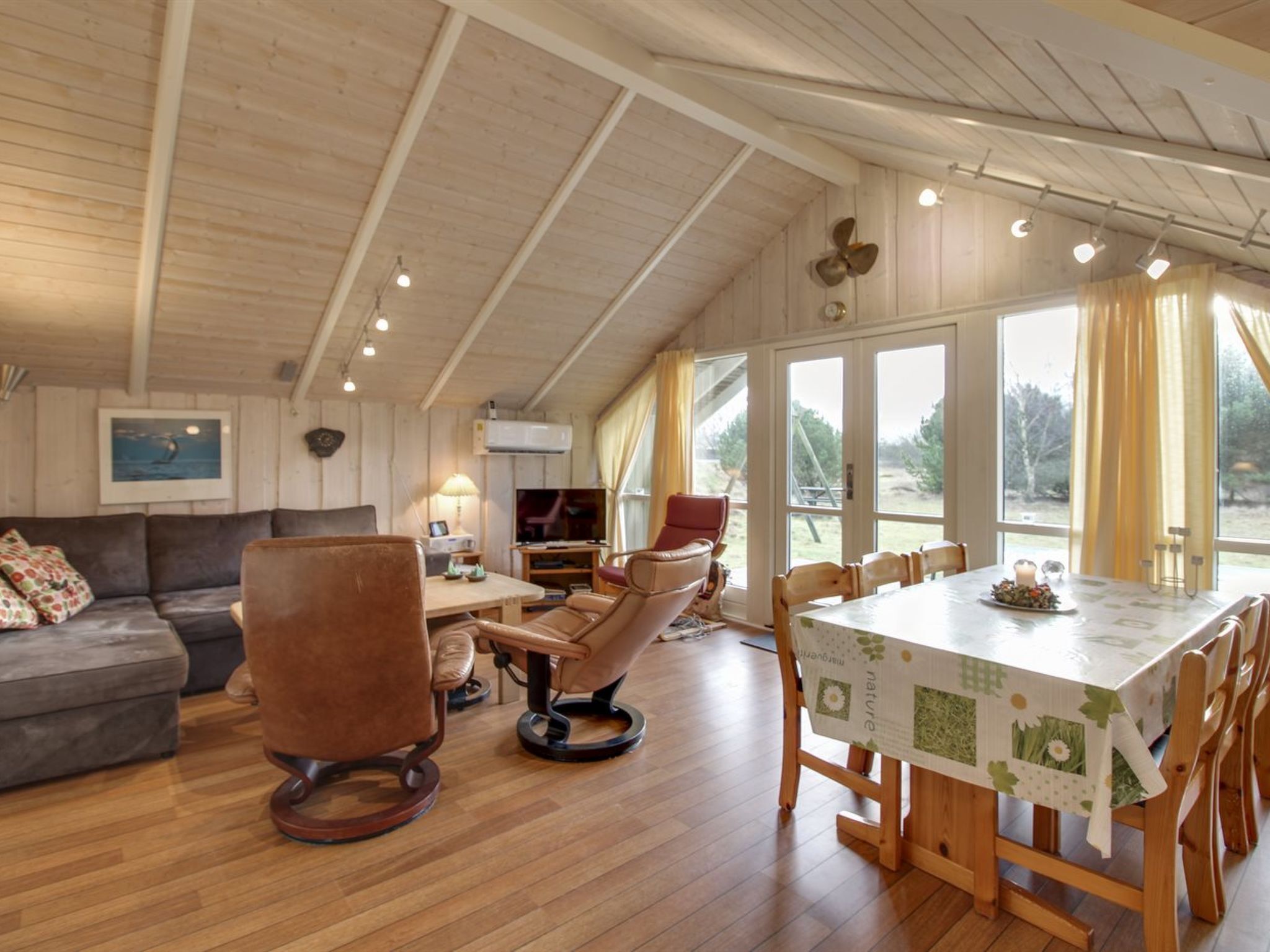 Ein geräumiger Wohnbereich mit Holzboden, einer Couch, einem braunen Ledersessel, einem Esstisch mit Tischdecke mit Naturmotiv und einem Fernseher.