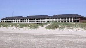 En lång, envåningsbyggnad med ett platt tak, belägen på en sandstrand med sanddyner i förgrunden.