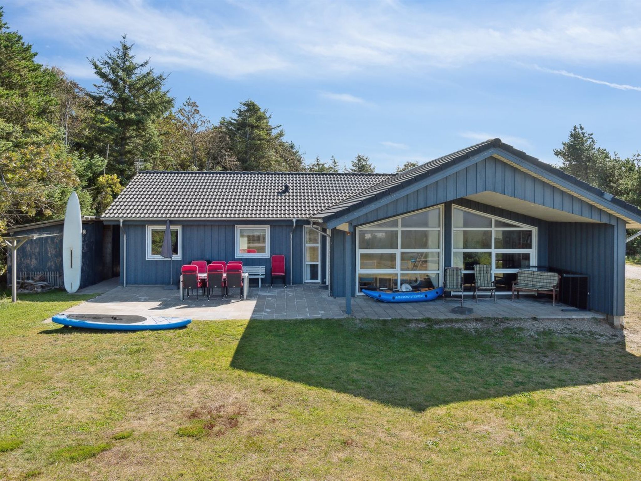 Une maison à deux étages avec un patio couvert, un coin repas avec des chaises rouges et une planche de surf appuyée contre la maison.