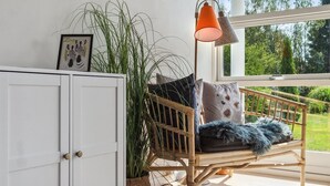 Een kamer met een rieten stoel, een witte kast, een grote potplant en uitzicht op groen buiten.