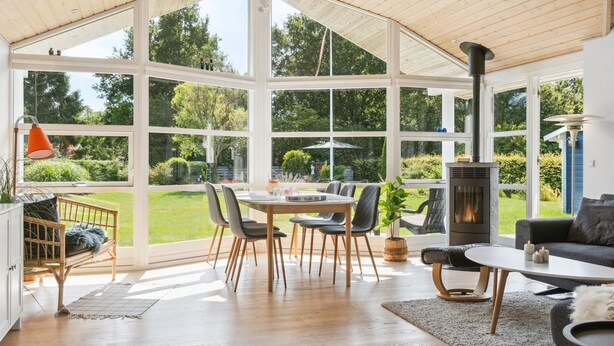 Een ruime kamer met een groot raam, houten plafond, een eettafel met stoelen, een bank en een open haard.