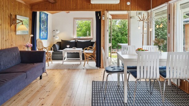 Een knus vertrek met een houten plafond, een bank, een eettafel met stoelen, en een raam met uitzicht op groen.