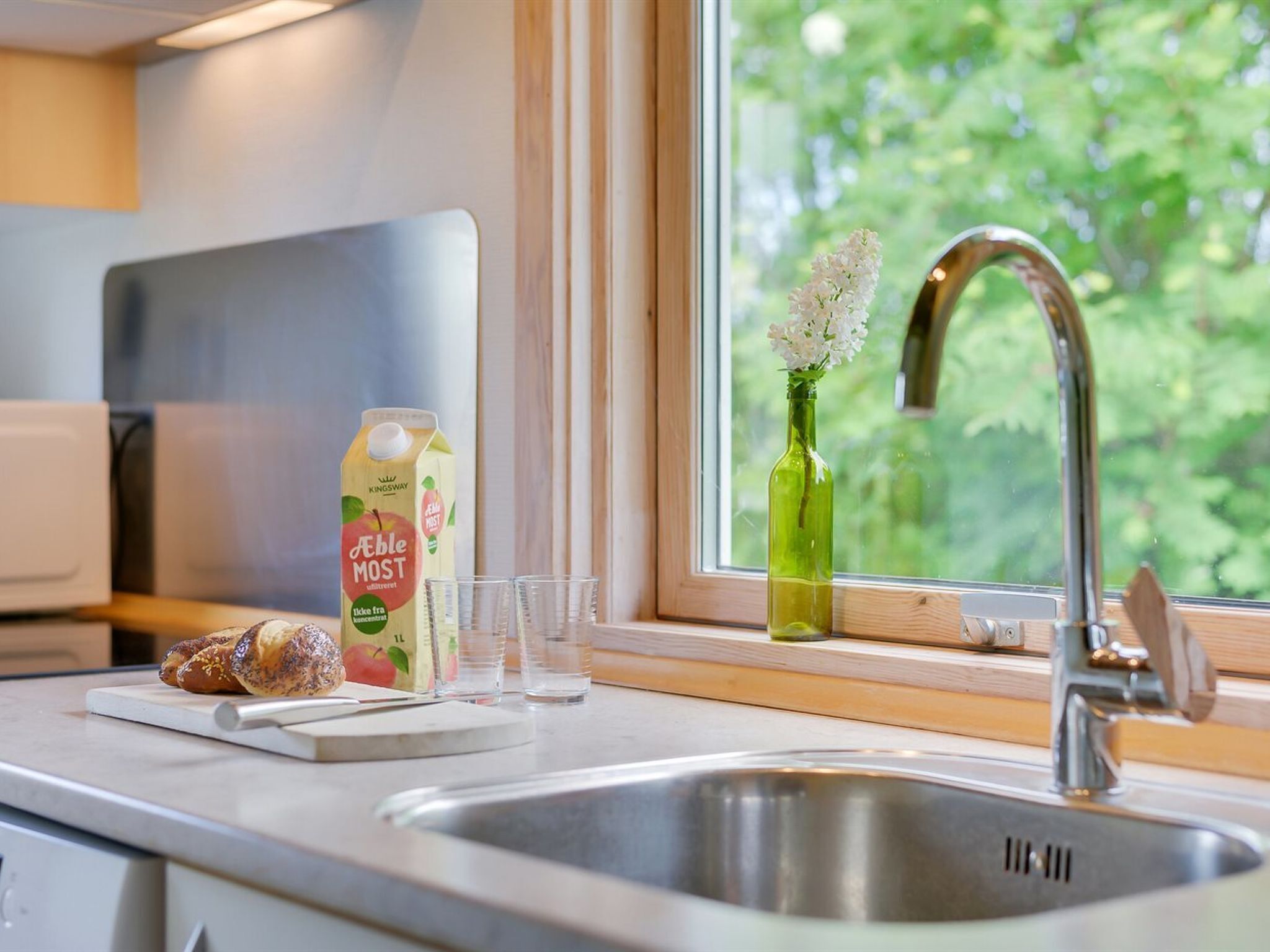Een keuken met een roestvrijstalen spoelbak, een raam met uitzicht op groen, een groene glazen fles met bloemen en een pak appelsap.
