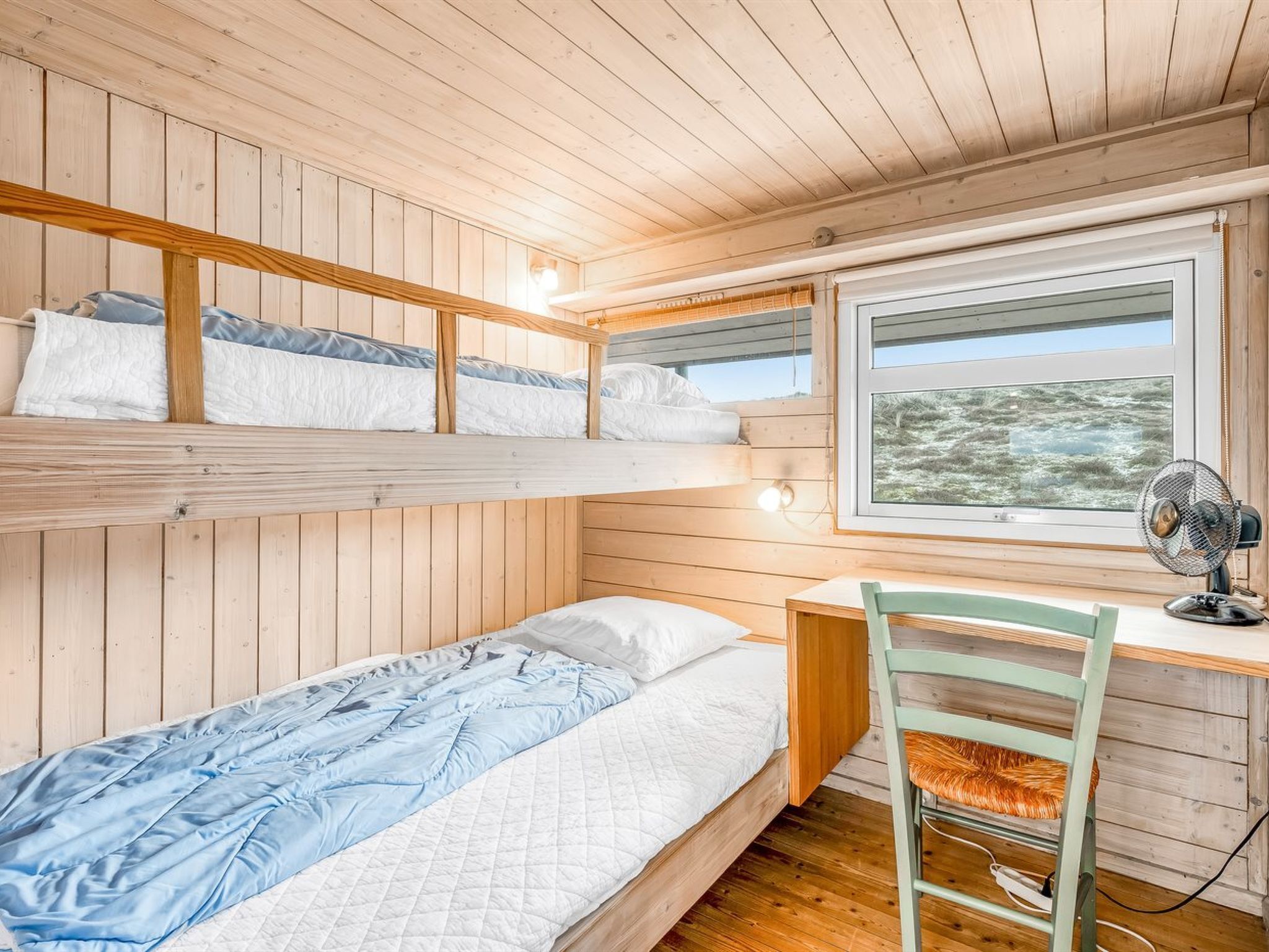 Une chambre avec un lit superposé, un bureau et une chaise, une fenêtre donnant sur l’extérieur et un ventilateur sur le bureau.