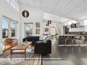 Ein großzügiger Wohnbereich mit Sofa, Couchtisch und Teppich. Es gibt eine Küchenzeile mit Kühlschrank, Spüle und Barhocker.