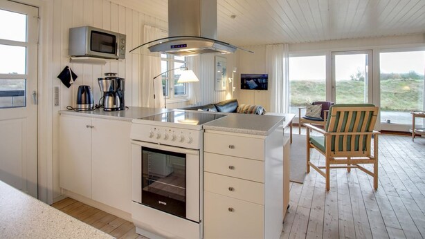 A modern kitchen with a built-in oven, microwave, and gas stove. There is a dining area with a wooden table and chairs, and a large window offering a view of the outdoors. - "Eikka" - 600m from the sea by Interhome (Fanø)