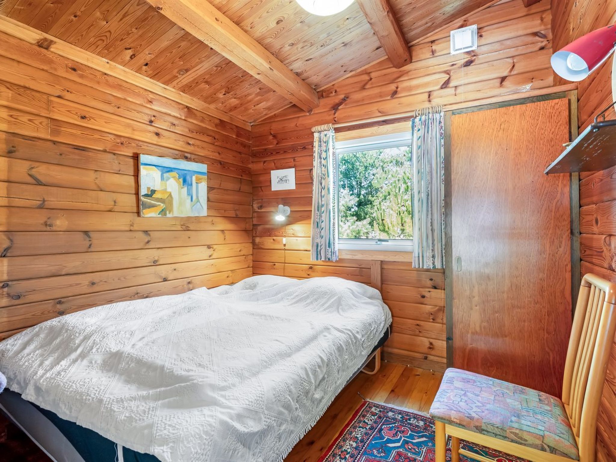Een houten kamer met een bed, een stoel, een raam met gordijnen en een schilderij aan de muur.