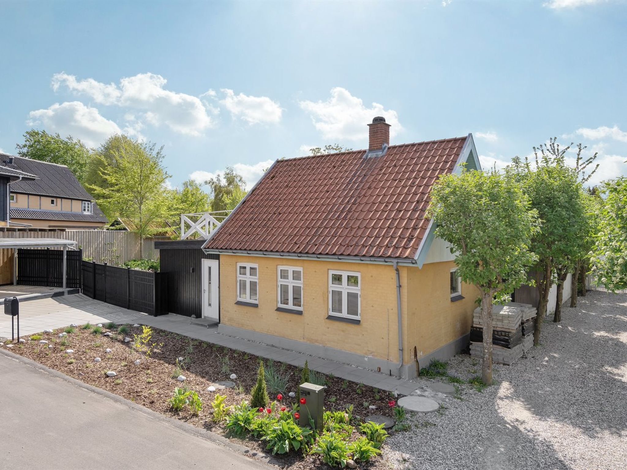 A single-story house with a brown tiled roof, a small garden with various plants and flowers, and a gravel driveway.