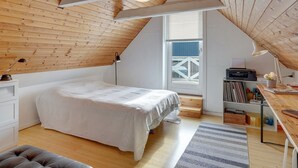 Ein gemütliches Schlafzimmer im Dachgeschoss mit schrägen Wänden, einem Bett, einem Schreibtisch mit Drucker und einem Fenster mit Blick auf einen weißen Zaun.