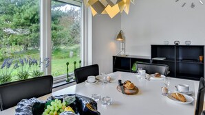 A modern dining area with a white table set for four, a chandelier, and a view of greenery outside.