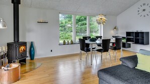 A modern living room with a wood-burning stove, dining area, and a large window with a view of greenery.