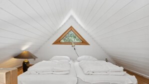 Une petite chambre mansardée de forme triangulaire, avec deux lits, une table de chevet, une lampe et une fenêtre donnant sur les arbres.