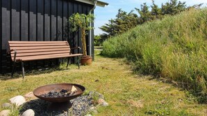 A wooden bench, a potted plant, and a fire pit in a grassy outdoor area with a building in the background.
