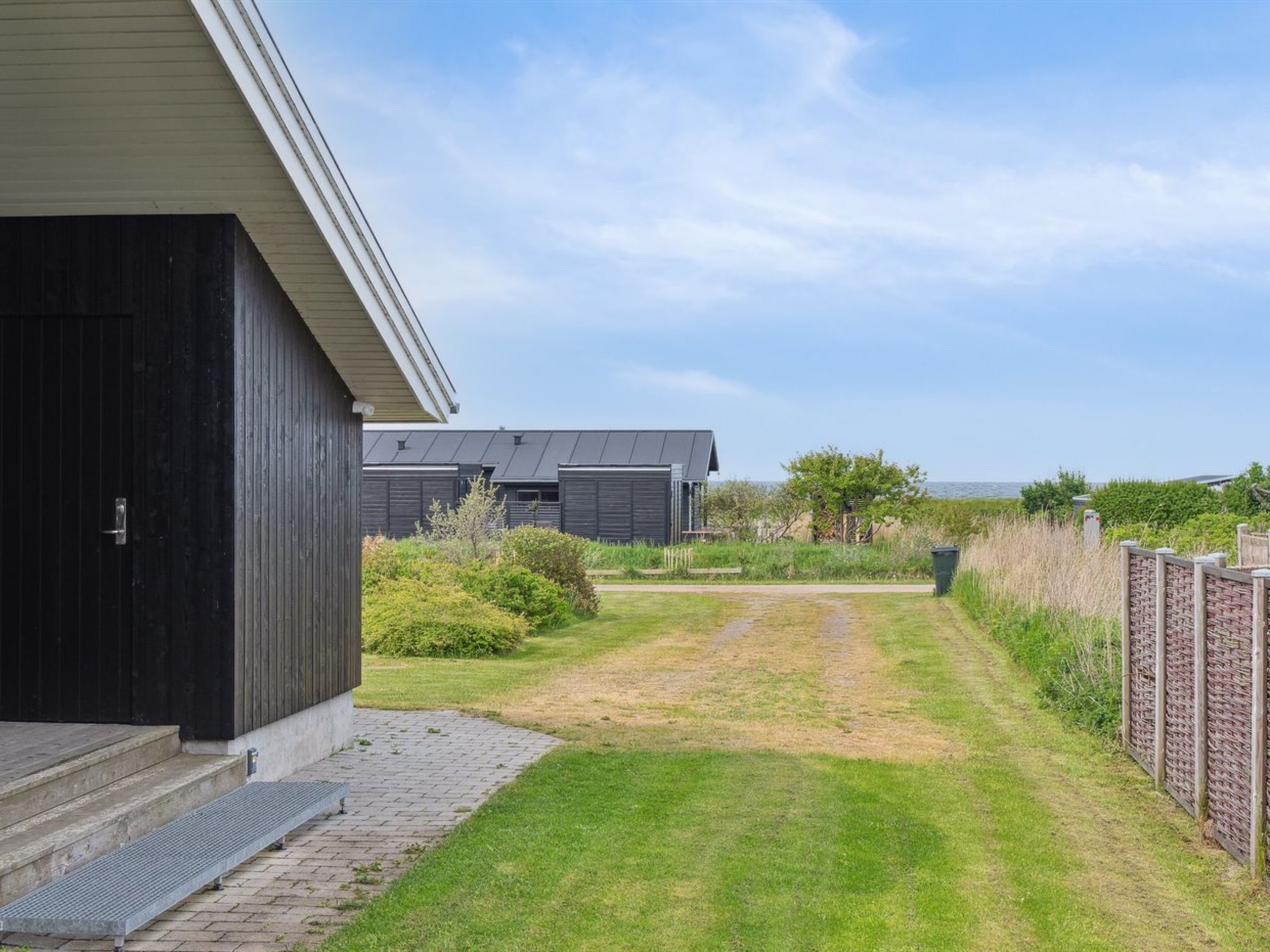 A modern building with a paved pathway leading to a grassy area, surrounded by vegetation and a body of water in the background.