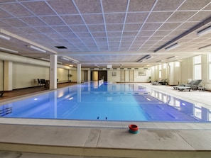 A large indoor swimming pool with a tiled ceiling, lounge chairs, and a small red object on the poolside.