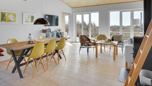 Un vaste espace dédié à la salle à manger, avec une table en bois et des chaises jaunes, un téléviseur fixé au mur et de grandes fenêtres offrant une vue sur l’extérieur.