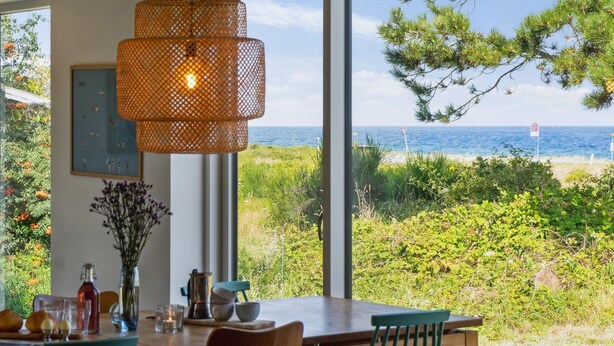 Un comedor con un ventanal que da a una playa, una lámpara colgante de mimbre, y una mesa dispuesta con vasos y un florero.
