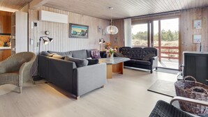 Ein modernes Wohnzimmer mit einer grauen Couch, einem Korbstuhl und einem schwarzen Ledersofa. Ein Holztisch mit einem Blumenvasen steht davor. Die Wände sind aus Holz und ein großes Fenster bietet Blick ins Freie.
