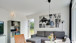 A modern living room with a grey sofa, a wooden chair, a small table with a vase, and a bookshelf.