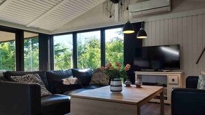 Un salon moderne avec un canapé, une table basse et un téléviseur. De grandes fenêtres offrent une vue sur la verdure environnante.