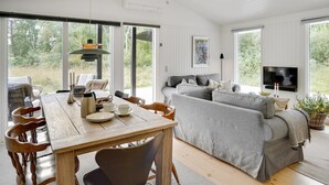 A spacious living area with a wooden dining table, grey sofa, and a TV mounted on the wall.
