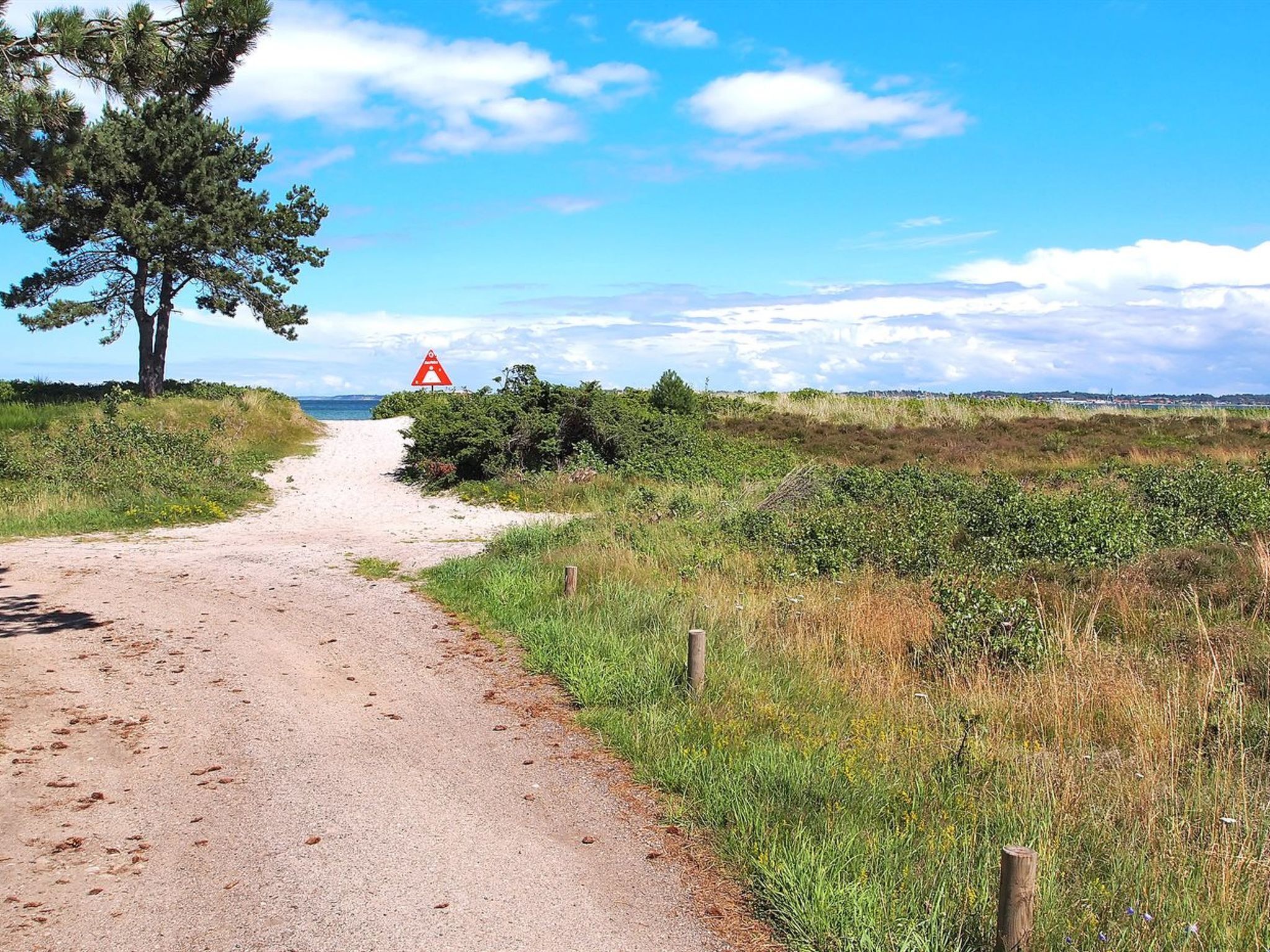 A gravel path leading to a coastal area with a warning sign, surrounded by greenery and a clear sky.
