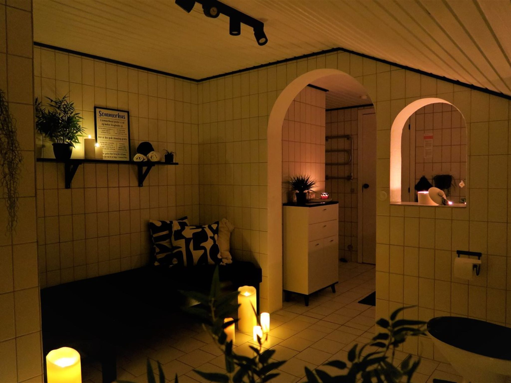 Une pièce faiblement éclairée, avec un sol carrelé, un canapé, une petite table sur laquelle se trouve une plante, et un miroir qui reflète une salle de bain.