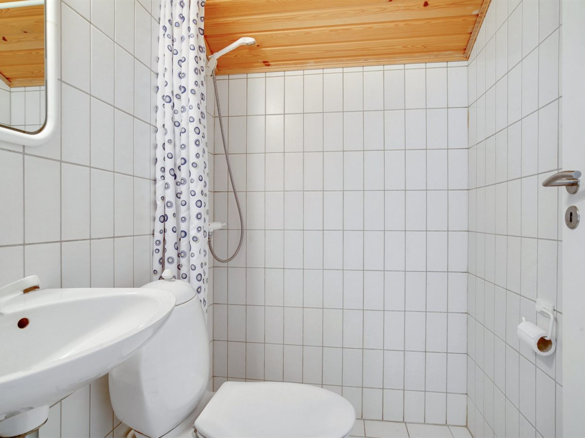 A small, clean bathroom with white tiles, a wooden ceiling, a shower with a curtain, a sink, and a toilet.