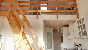 A loft bedroom with a wooden staircase, a kitchen with white cabinets, and a door leading to another room.