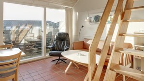 A modern living room with a large window, a black leather chair, a wooden table, and a wooden staircase.