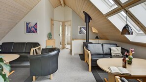 A modern loft-style living room with a sloped ceiling, wooden beams, and skylights. There are two black leather sofas, a wooden coffee table, and a small dining table with a book and a bottle.