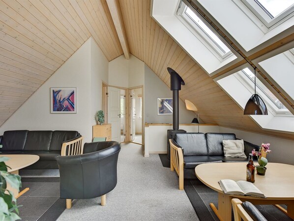 A modern loft-style living room with a sloped ceiling, wooden beams, and skylights. There are two black leather sofas, a wooden coffee table, and a small dining table with a book and a bottle.
