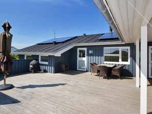 Un espace extérieur abrité doté d’une terrasse en bois, d’un barbecue et d’un coin salon avec des fauteuils en osier.