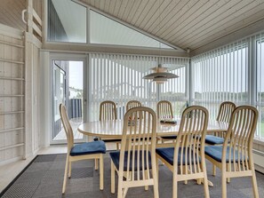 Un coin repas avec une table ronde et six chaises, de grandes fenêtres équipées de stores verticaux et un ventilateur de plafond.