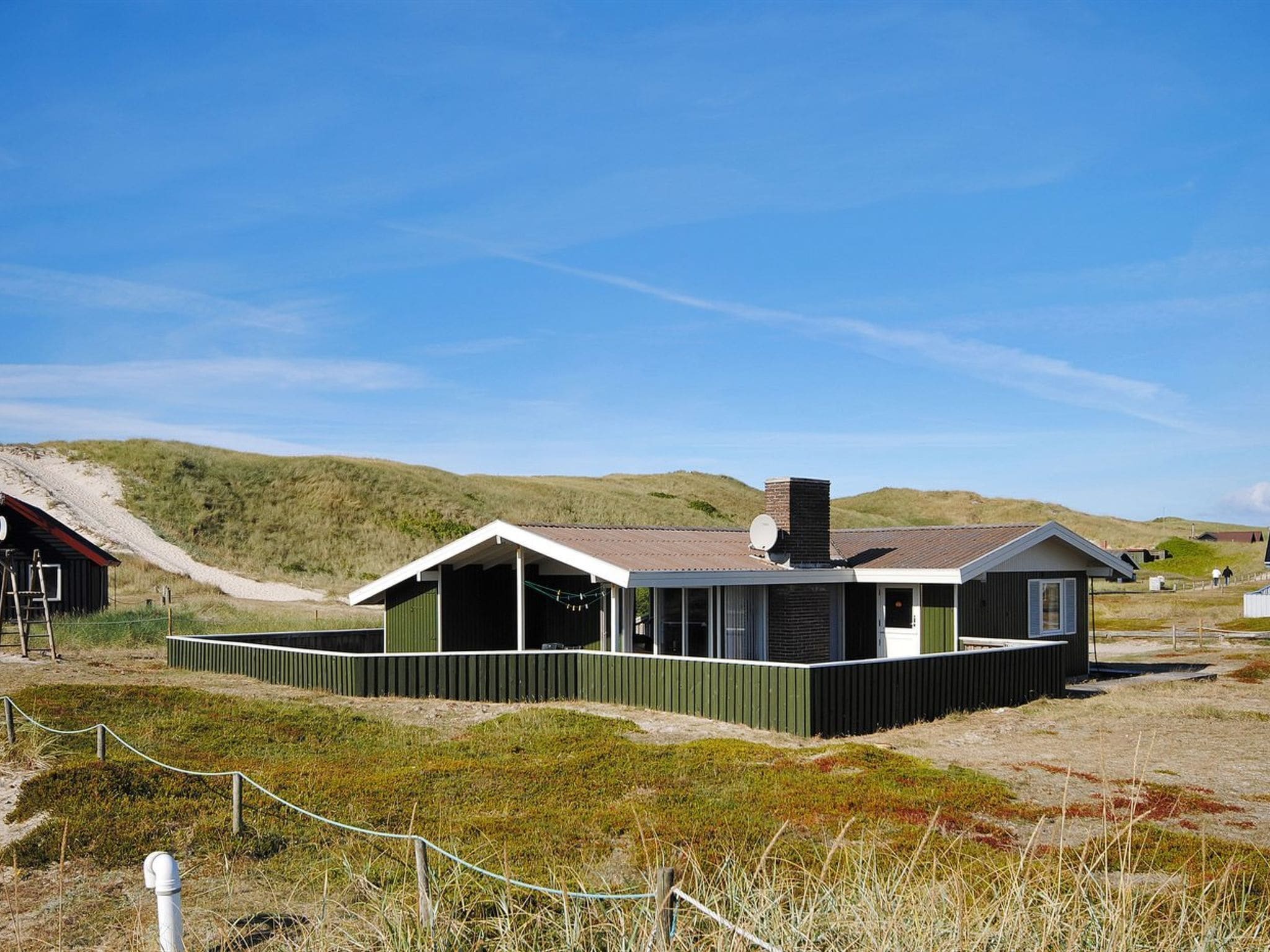 A modern building with a flat roof and a chimney, surrounded by a fenced area with grassy terrain and hills in the background.