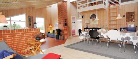 A spacious living area with a high ceiling, wooden floors, a brick fireplace, and a dining area with a table and chairs.