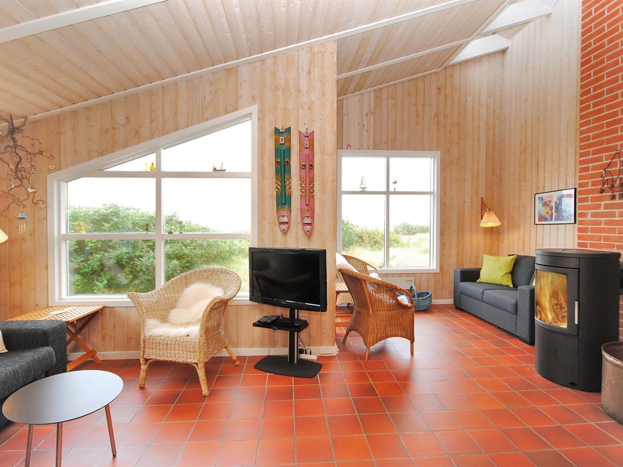 A cozy living room with a TV, wicker chairs, a fireplace, and large windows offering a view of greenery.