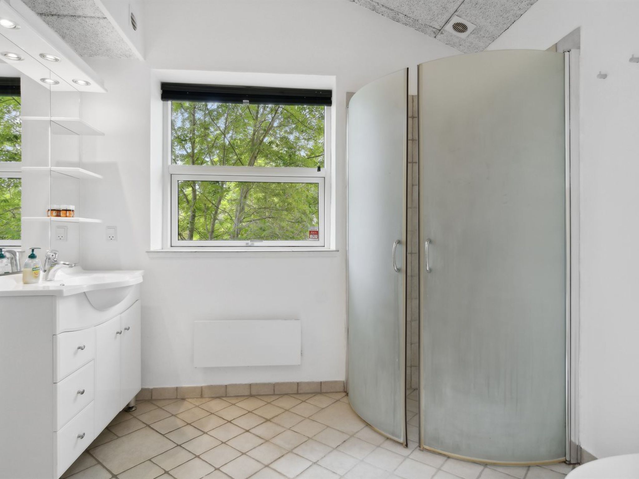 Un baño moderno con un tocador blanco, un ventanal con vistas a árboles, una ducha con puertas de vidrio esmerilado y un suelo de baldosas.