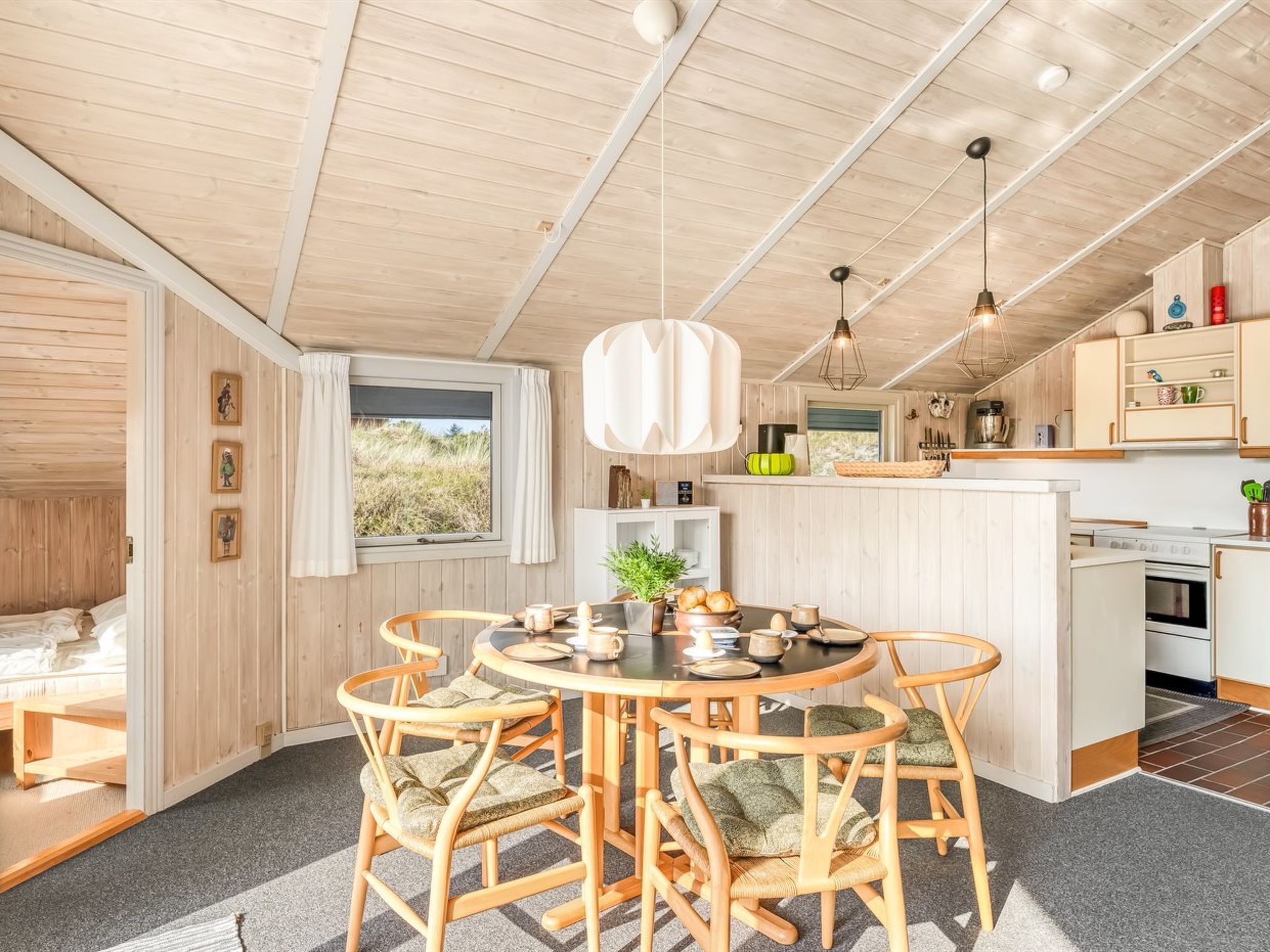 A cozy cabin interior with a round dining table, wooden chairs, a kitchen area, and a large window offering a view of the outdoors.