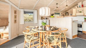 Interior acogedor de una cabaña con una mesa redonda para comer, sillas de madera, una zona de cocina y un amplio ventanal con vistas al exterior.