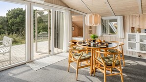 Un interior moderno con zona de comedor que incluye una mesa redonda y sillas de mimbre, un armario blanco con puertas de cristal y un gran ventanal corredero que da a una terraza exterior.