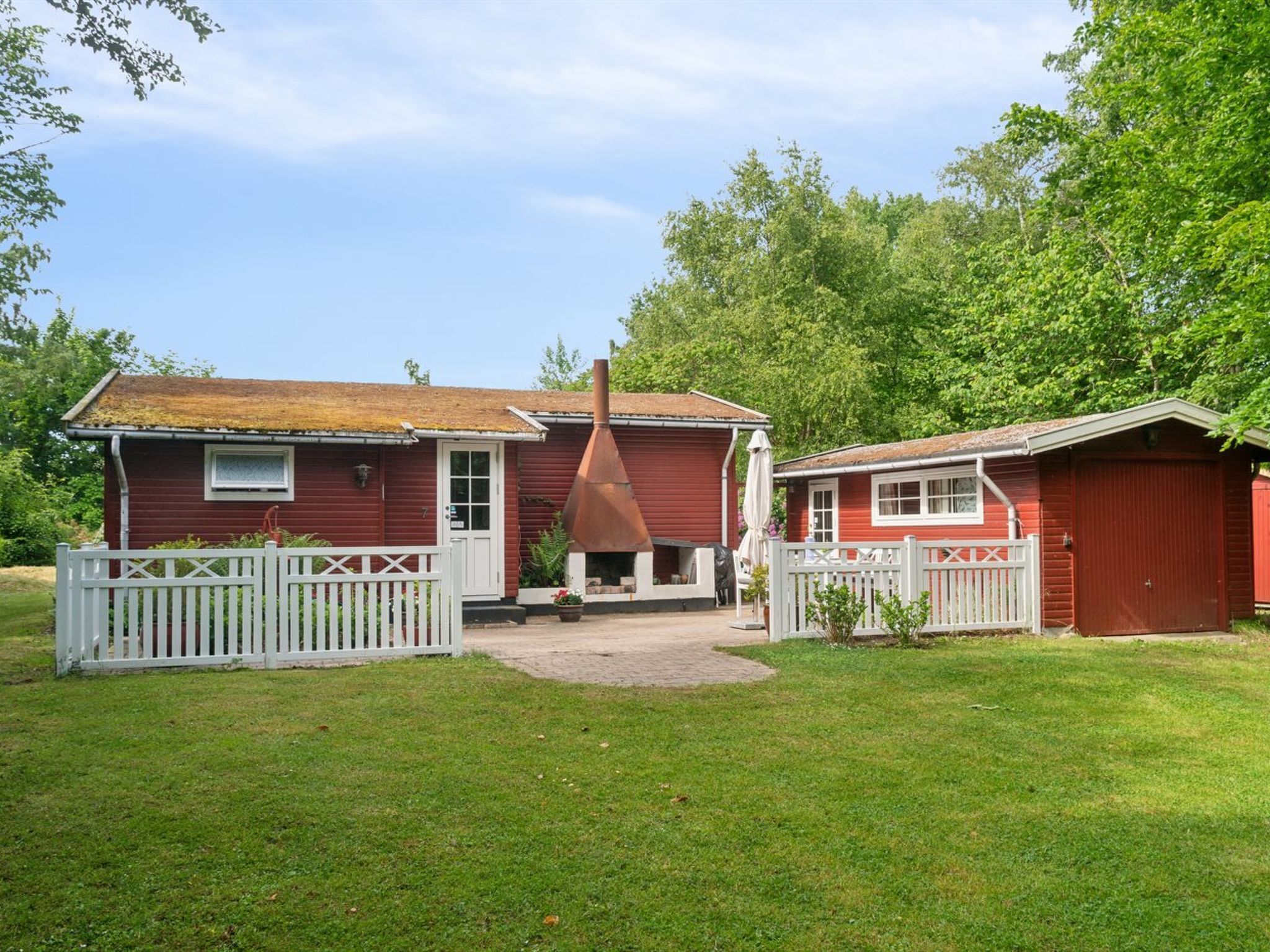 A red wooden house with a chimney, a white picket fence, and a small shed.