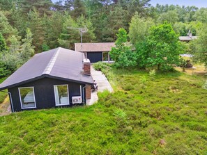 Ein Haus mit dunkler Fassade, einem Ziegeldach und einem Schornstein. Es ist von üppiger Vegetation und Bäumen umgeben.