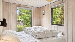 A bedroom with a wooden ceiling, two beds with white bedding, a black lamp, and a large window with a view of greenery.