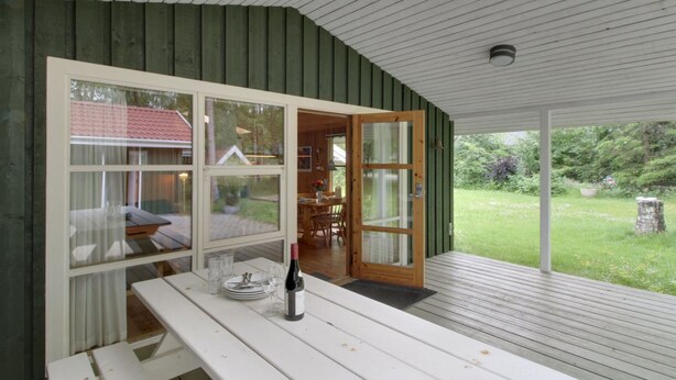 A covered outdoor dining area with a wooden table and benches, a bottle of wine, and a view of a garden and house through large windows.