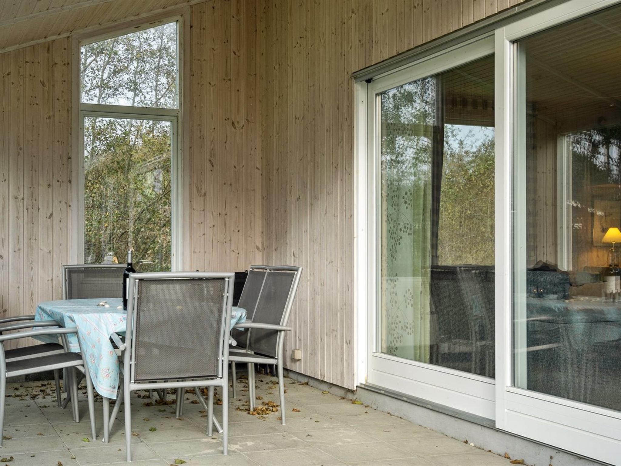 A covered outdoor seating area with a table and chairs, large windows, and a view of trees.